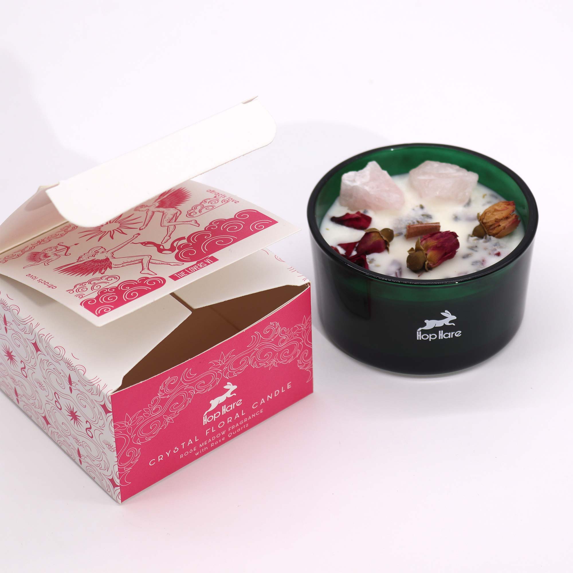 Vela aromática 'Floral Rose Meadow' con cristales de cuarzo rosa en un recipiente de vidrio rosado, sobre su caja de embalaje con un diseño floral y la promesa de una fragancia a rosas. La caja abierta revela la vela, ideal para regalo, relajación y decoración con un toque romántico.