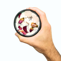 Mano sosteniendo una vela aromática 'Floral Rose Meadow' de cera blanca con cristales de cuarzo rosa, en un recipiente de vidrio rosado. La vela encendida emite un brillo cálido, ideal para momentos de meditación, autocuidado y para disfrutar de la fragancia a prado de rosas